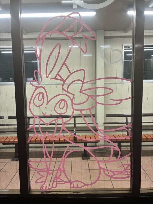 南大沢駅ホーム待合室のガラスに描かれたニンフィアの近景。リボン状の耳と長い尻尾の輪郭がピンクの線で描かれ、スタンプラリーの担当ポケモンとして待合室を飾っている
