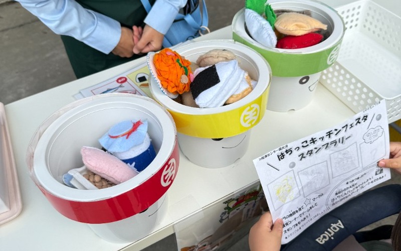 はちっこキッチンフェスタのスタンプラリー台紙と、赤黄緑の容器に入った食べ物のぬいぐるみ