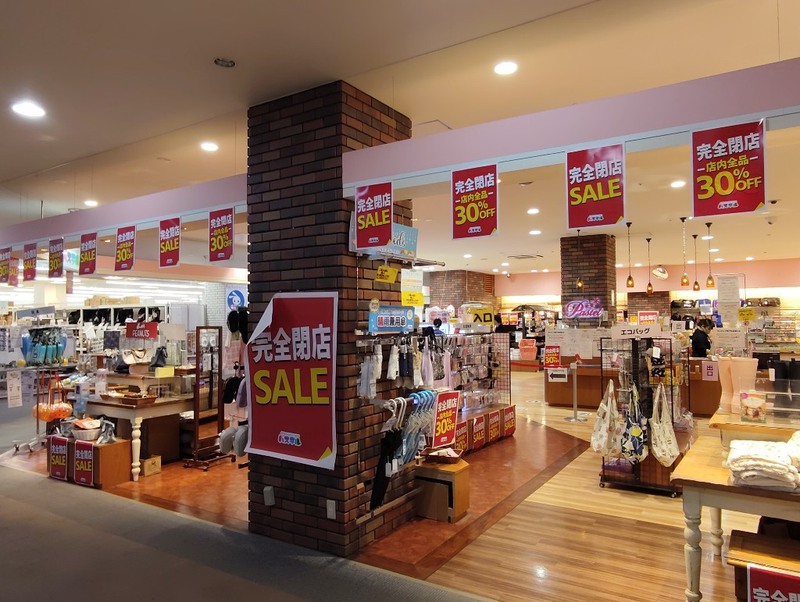 レンガの柱に完全閉店SALEのポスターが貼られたパステル多摩センター店の入口。晴雨兼用傘やエコバッグが並ぶ