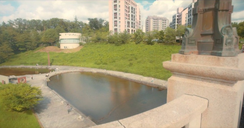 長池見附橋の親柱と欄干越しに、姿池と芝生の斜面、周辺のマンションを見下ろすカット