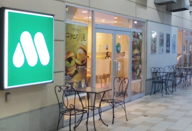屋外側のモスバーガー クロスガーデン多摩店の入口。緑のMマークの看板と今年のモスはコクとろ月見のポスター、アイアンのテラス席